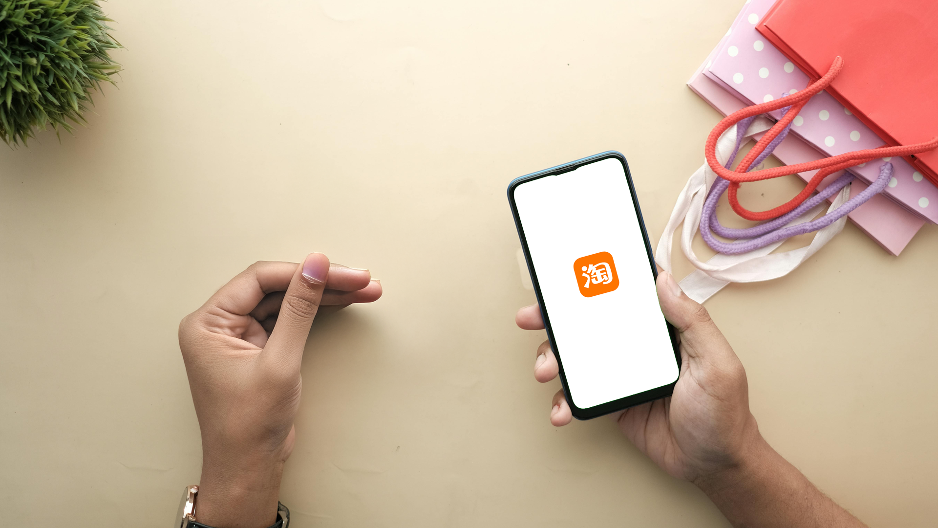 Gambar orang memegang ponsel yang menampilkan logo Taobao di tangan kanannya, di sekitar terdapat 3 paper bag dan bunga hias